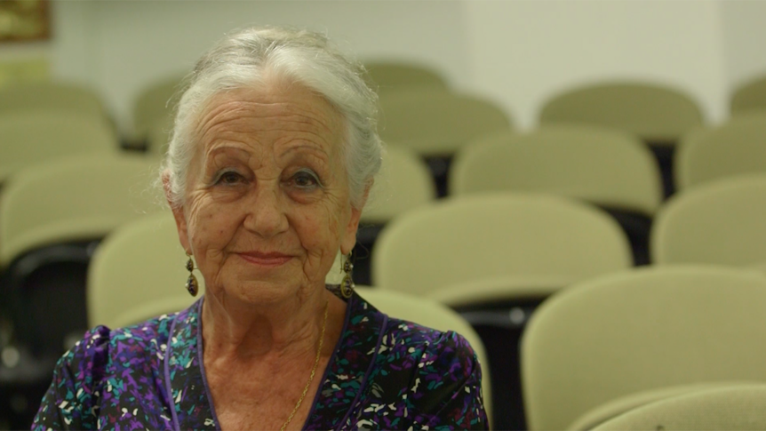 An old lady with grey hair is smiling in a museum surrounded by chairs.