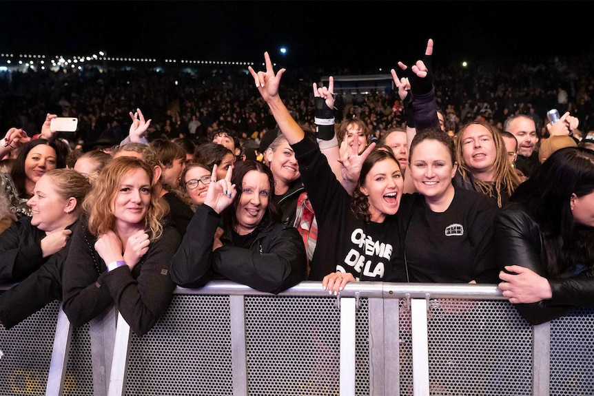 A crowd of people in the front row of a festival. Two women hold up the devil horns hand sign while grinning.