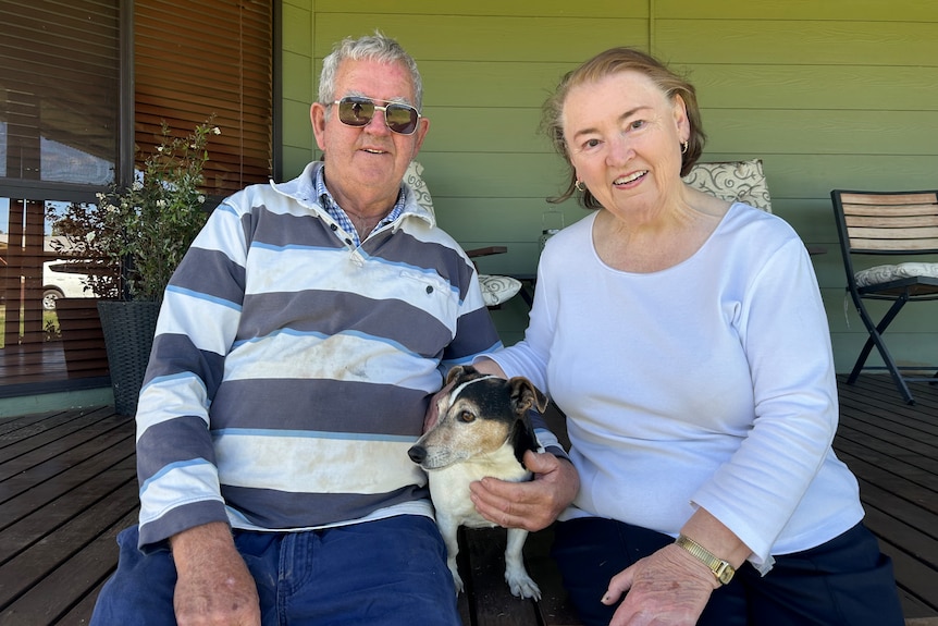 A smiling man and a woman sit on a porch with their dog between them.