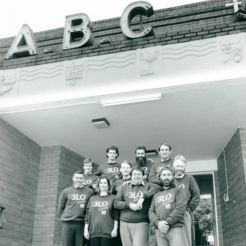 Black and white photo of ten people standing on steps, wearing 3LO jumpers, outside old ABC building.