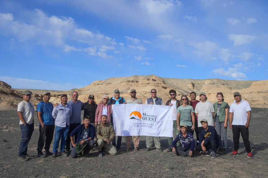 A group photo of palaeontologist in the Gobi Desert, holding a banner, blue skies, clouds.