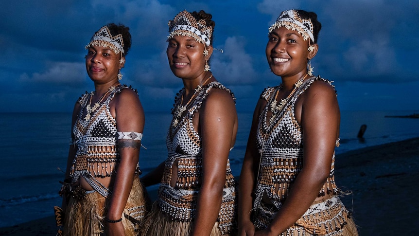 Three dancers from the Solomon Islands stand on the beach, dressed in traditional costumes made from shells and grass.