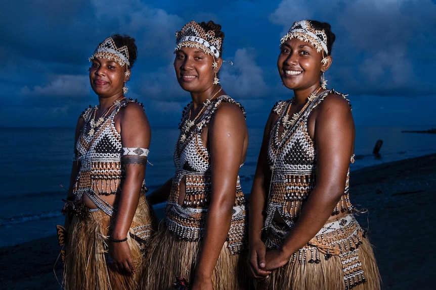 Three dancers from the Solomon Islands stand on the beach, dressed in traditional costumes made from shells and grass.