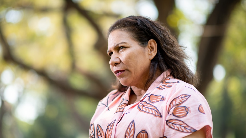 A woman looks at the camera with a stern expression. She wears a pink shirt.