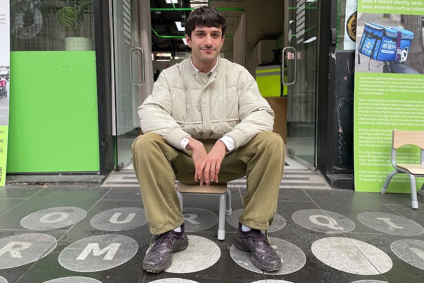 Andrew Copolov sits on small chair outside shop doors on the street