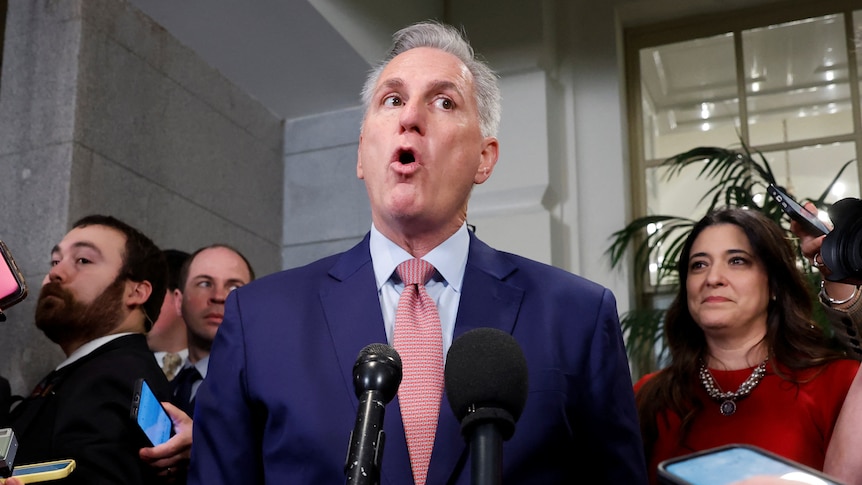Kevin McCarthy gives a defiant press conference, wearing a blue suit and pink tie, his mouth shaped like an "O"