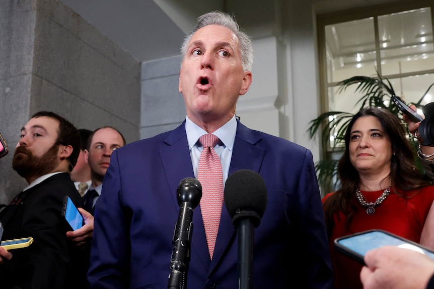 Kevin McCarthy gives a defiant press conference, wearing a blue suit and pink tie, his mouth shaped like an "O"