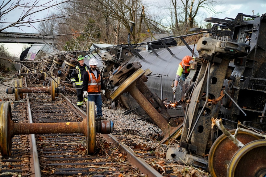 Men working on a derailed train on its side by railway tracks.