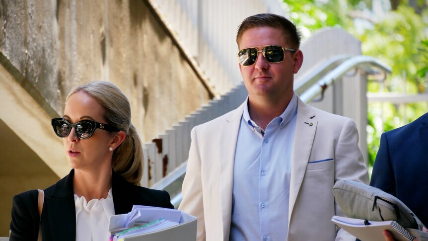A woman (left) and man (right) dressed in corporate attire, wearing sunglasses, walk outside.