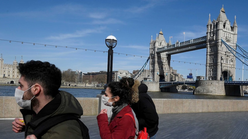 A man and a woman wearing face masks stand in front of Tower Bridge in London on a clear blue sky day.