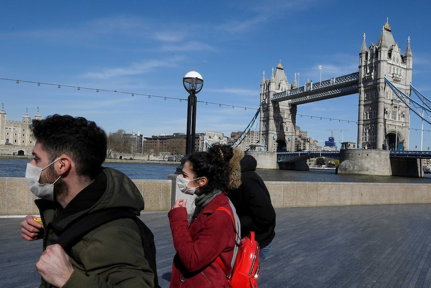 A man and a woman wearing face masks stand in front of Tower Bridge in London on a clear blue sky day.