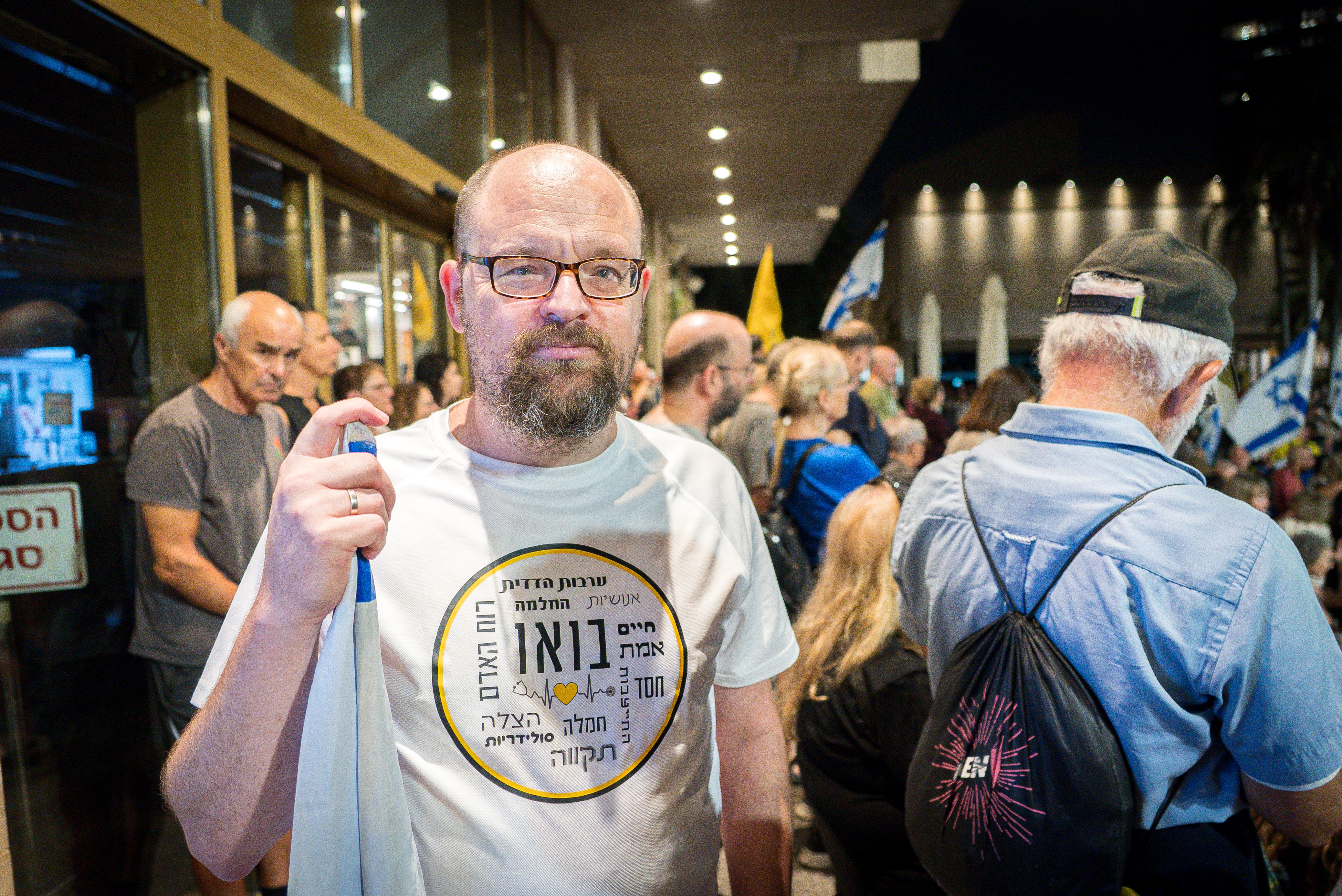 A man with glasses stands in a crowd, wearing a white tee-shirt and holding a rolled-up flag.