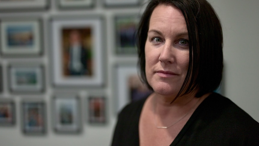 A mother stands in front of a wall of photos of her son.