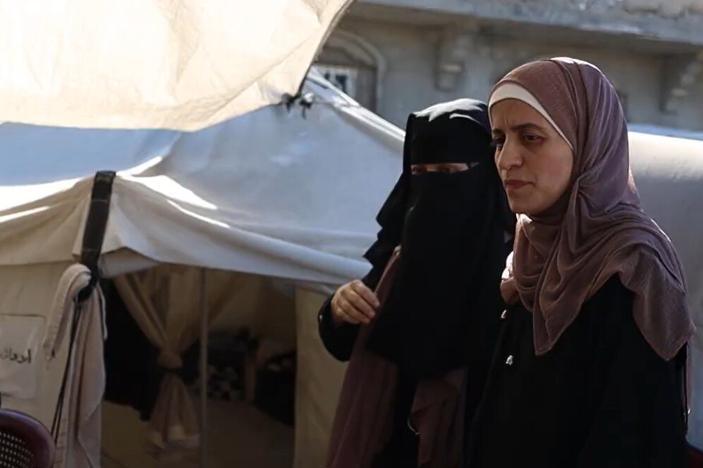 Mervat Sarhan, dressed in traditional Palestinian clothes, stands next to a woman in a camp.