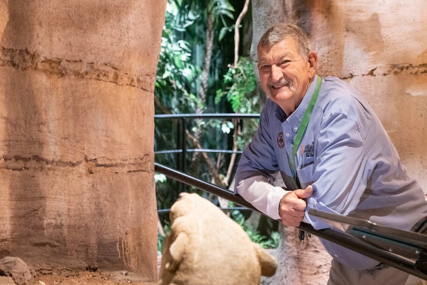 An older gentleman leans against a museum exhibit barrier, smiling.