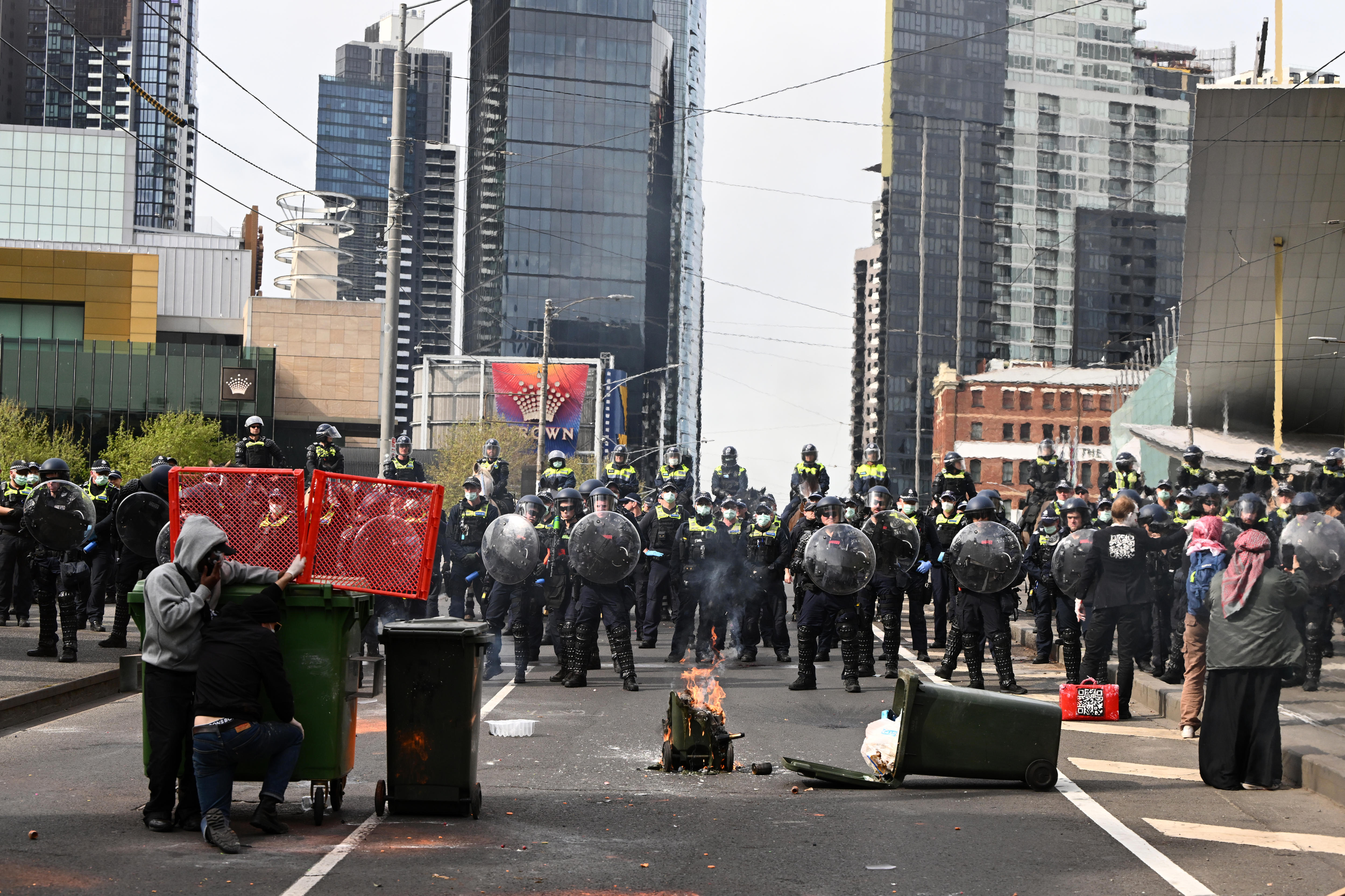 A fire burns in the middle of a road as police stand in a line with shields up.