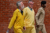 Two white men wear yellow jumpsuits as they walk past a brick wall, escorted by an official in a brown suit