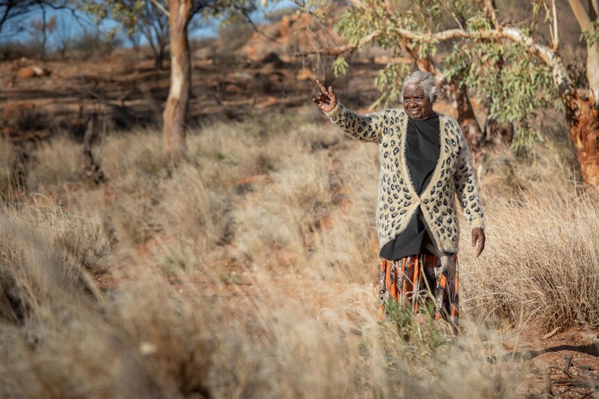 An older Aboriginal woman with grey hair, the artist Betty Muffler, stands in the bush with one arm raised to the sky