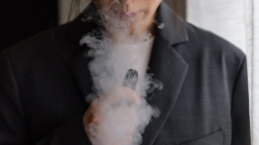 a close-up image of a woman's hand holding a vape. smoke can be seen coming from her nose and moutn