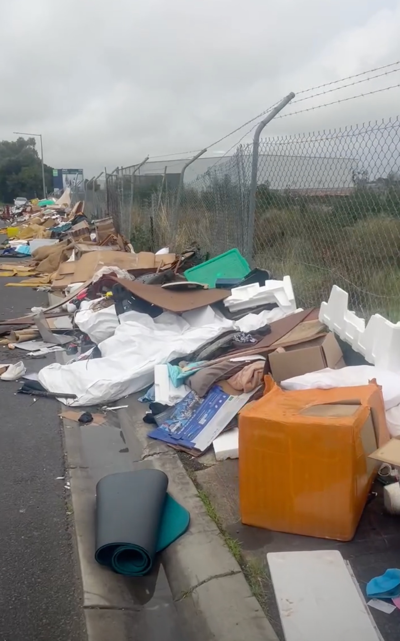 Piles of rubbish along a fence, in a carpark, alongside a road. 