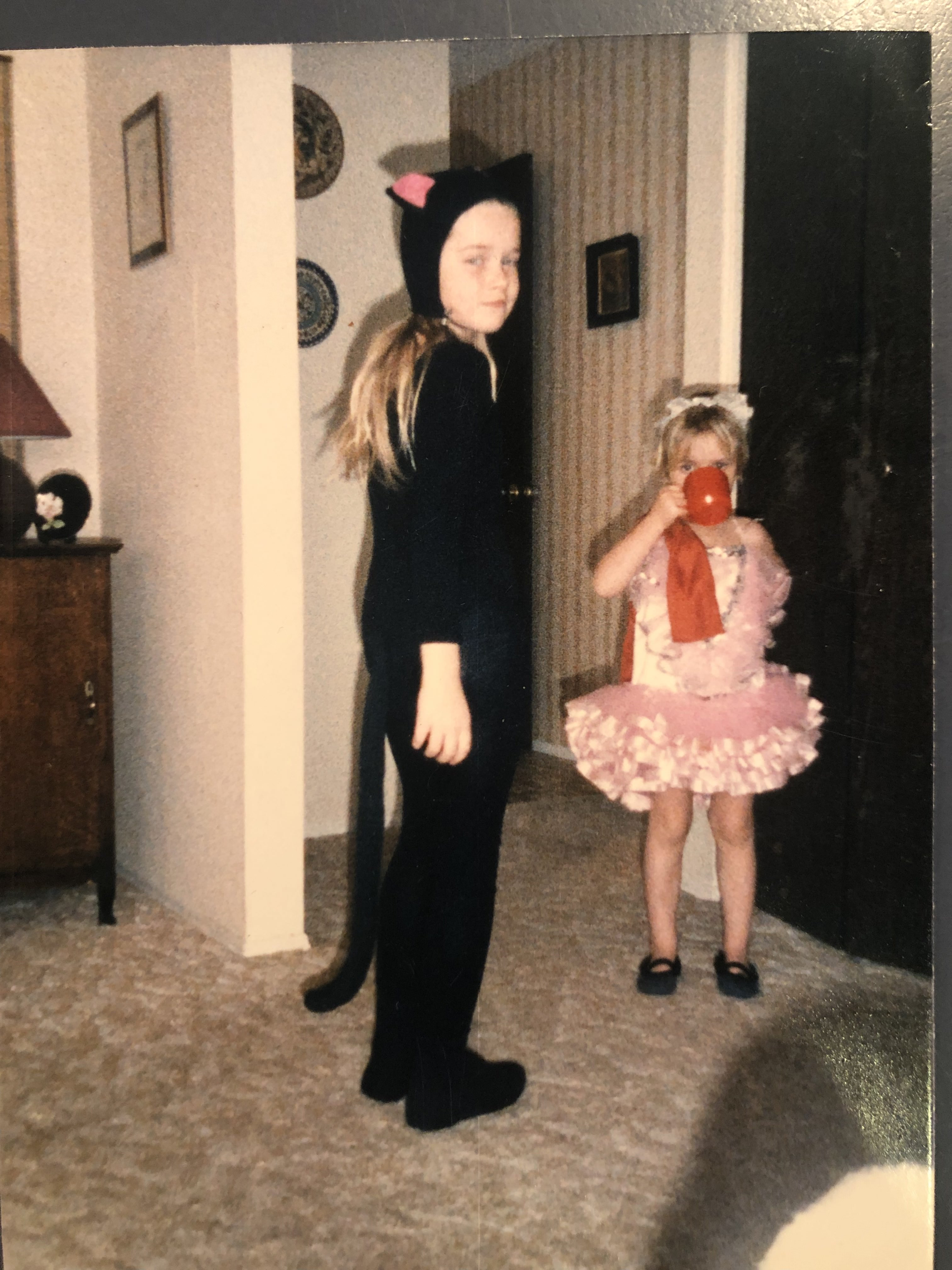 Young blond white girl dressed in a black cat costume stands in living room beside even smaller girl in a pink tutu.
