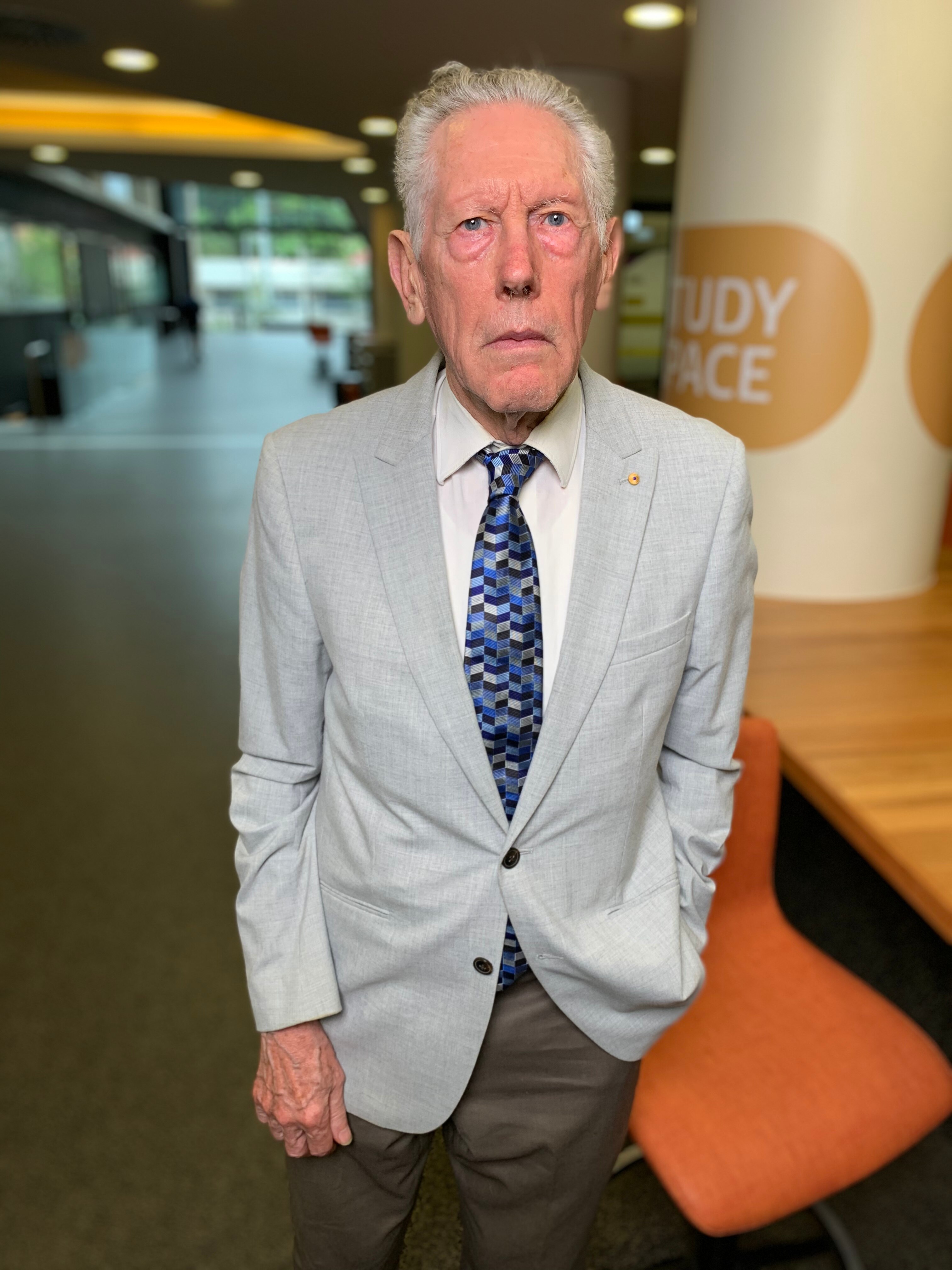 Older man in a light grey suit jacket and grey pants with a blue tie.