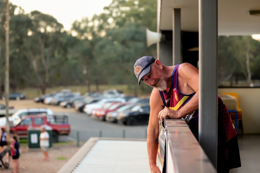 Older man wearing cap, singlet and shorts stands leans over ledge fixing a hanging sign