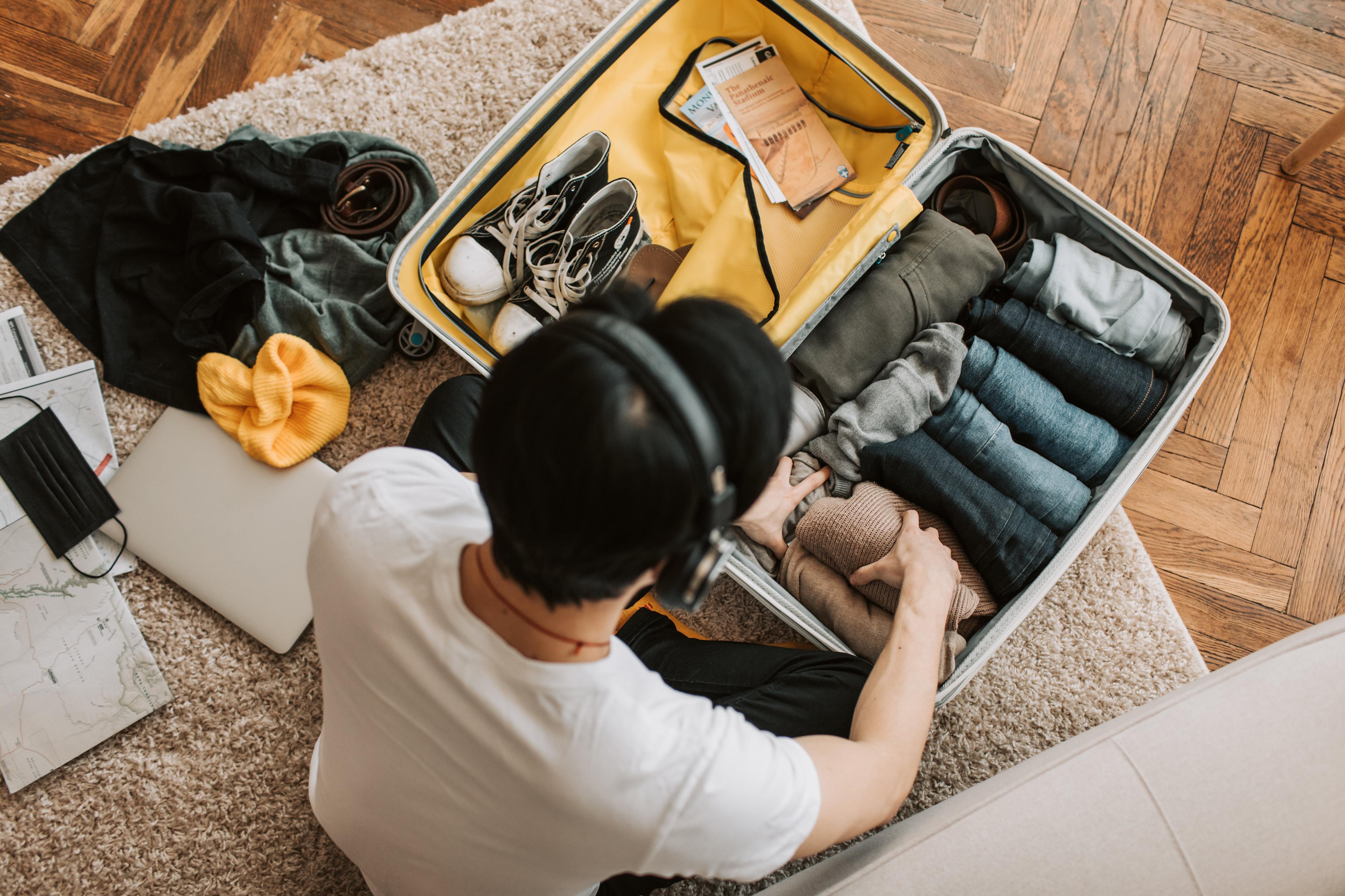 A man sits on a floor, wearing headphones, and packing a suitcases with clothes and Converse sneakers