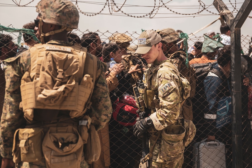 A US soldier stands guard in front of a chain link fence, behind which queue dozens of people at Kabul's airport