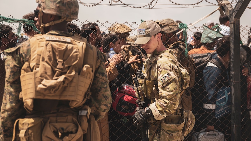 A US soldier stands guard in front of a chain link fence, behind which queue dozens of people at Kabul's airport