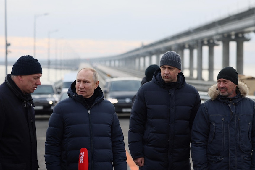 Four older men in dark blue winter clothing stand on a long concrete bridge in front of a selection of cars.