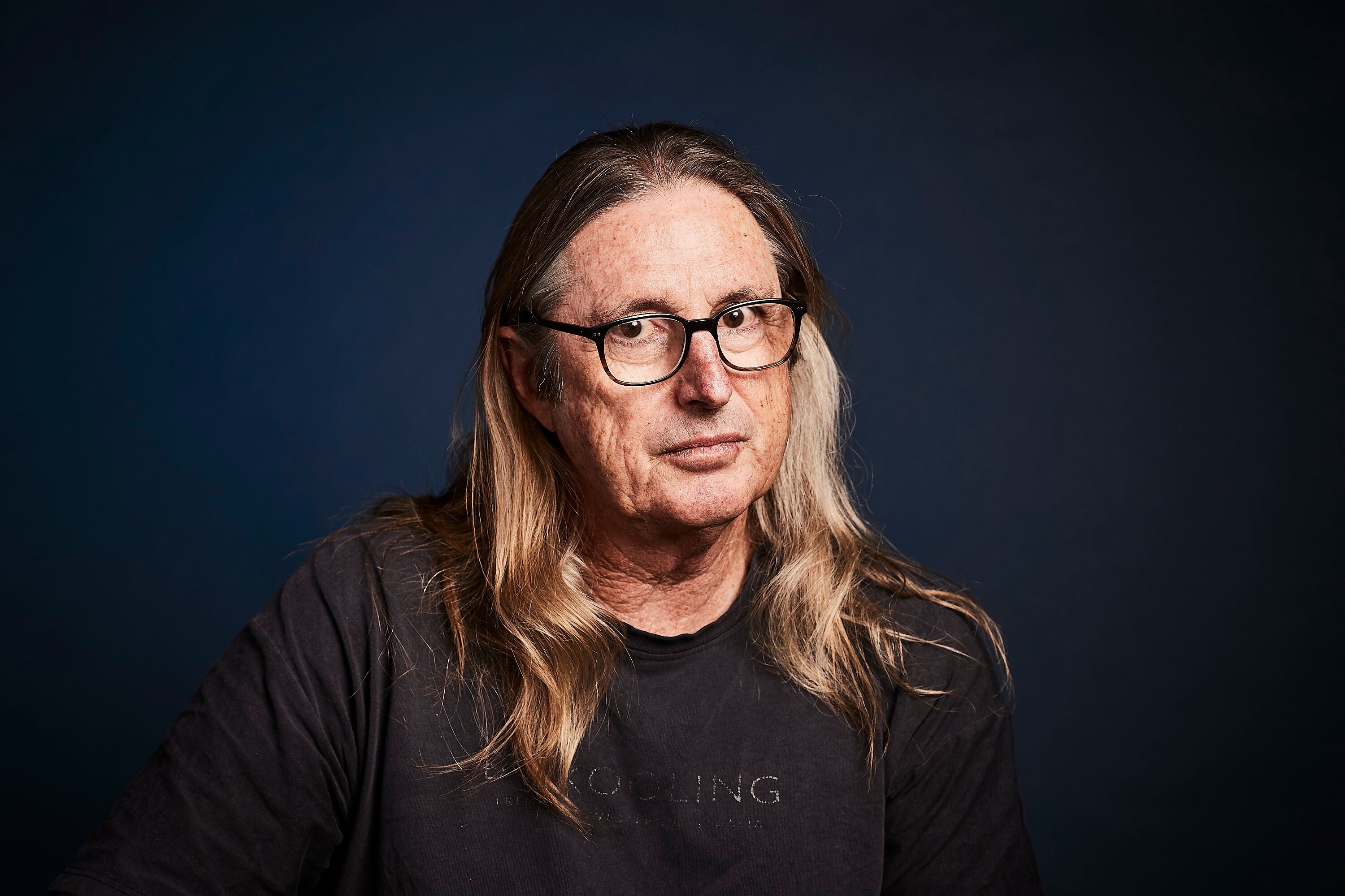 A man with long sandy-coloured hair wearing thick-framed glasses looks directly at the camera without smiling