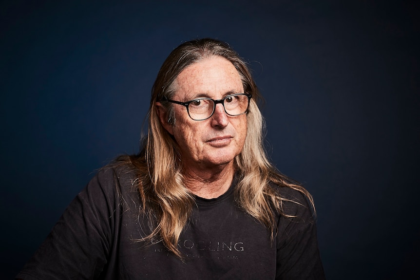 A man with long sandy-colored hair wearing thick-framed glasses looks directly at the camera without smiling.