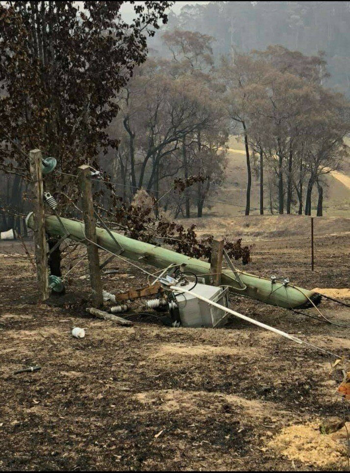 A broken power pole on the ground with burnt trees in the background.