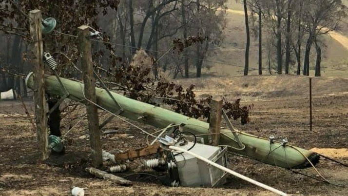 A broken power pole on the ground with burnt trees in the background.