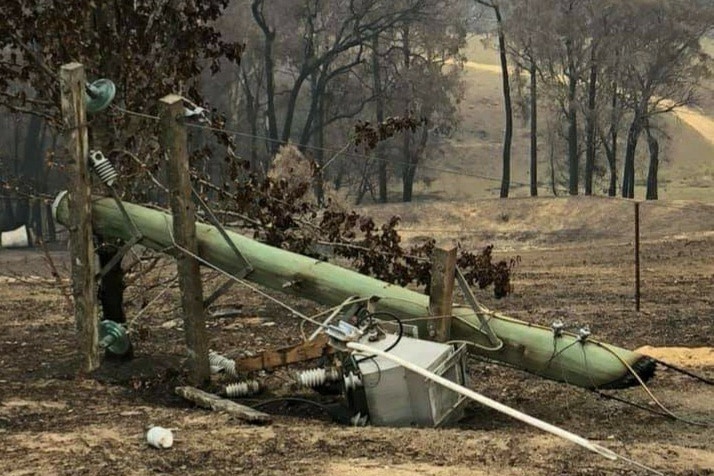 A broken power pole on the ground with burnt trees in the background.