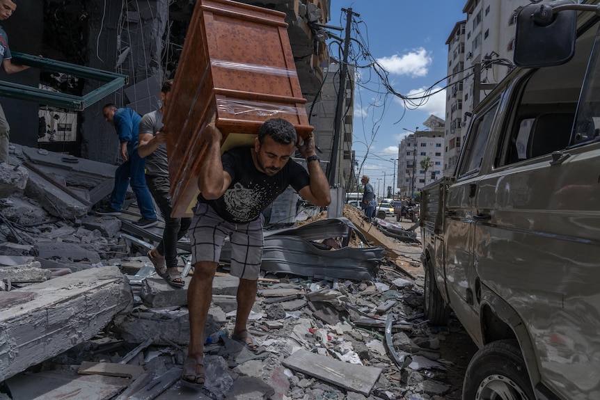 A young Arab man hoists a chest of drawers on his back as he walks through rubble 