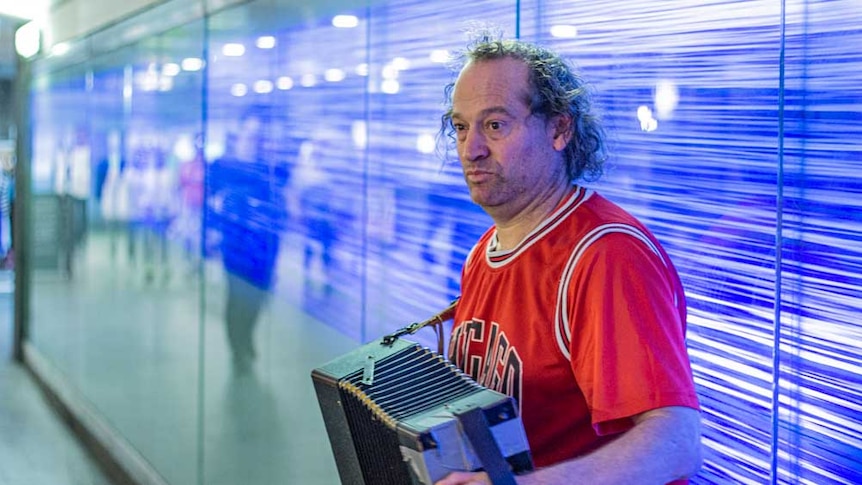 A man in a red basketball shirt plays accordion in a busy city tunnel against a blue background
