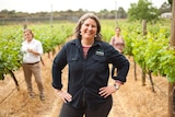 A woman stands, hands on hips, smiling at the camera between vine rows, with women checking the vines in the distance