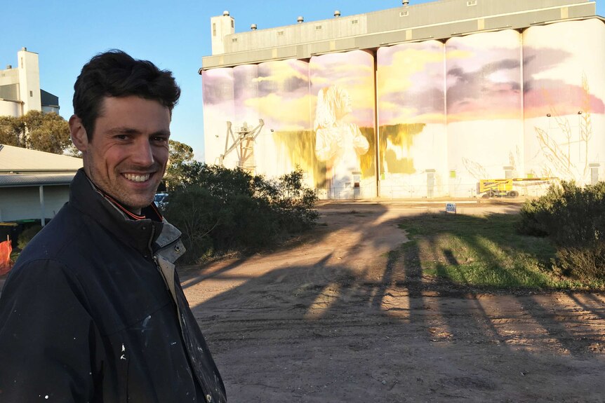 A man smiling with half-finished mural paintings on silos behind him