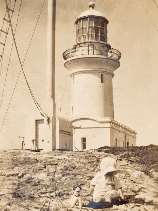An historic black and white photo shows a toddler sitting on rocks with a soft toy in front of a lighthouse.