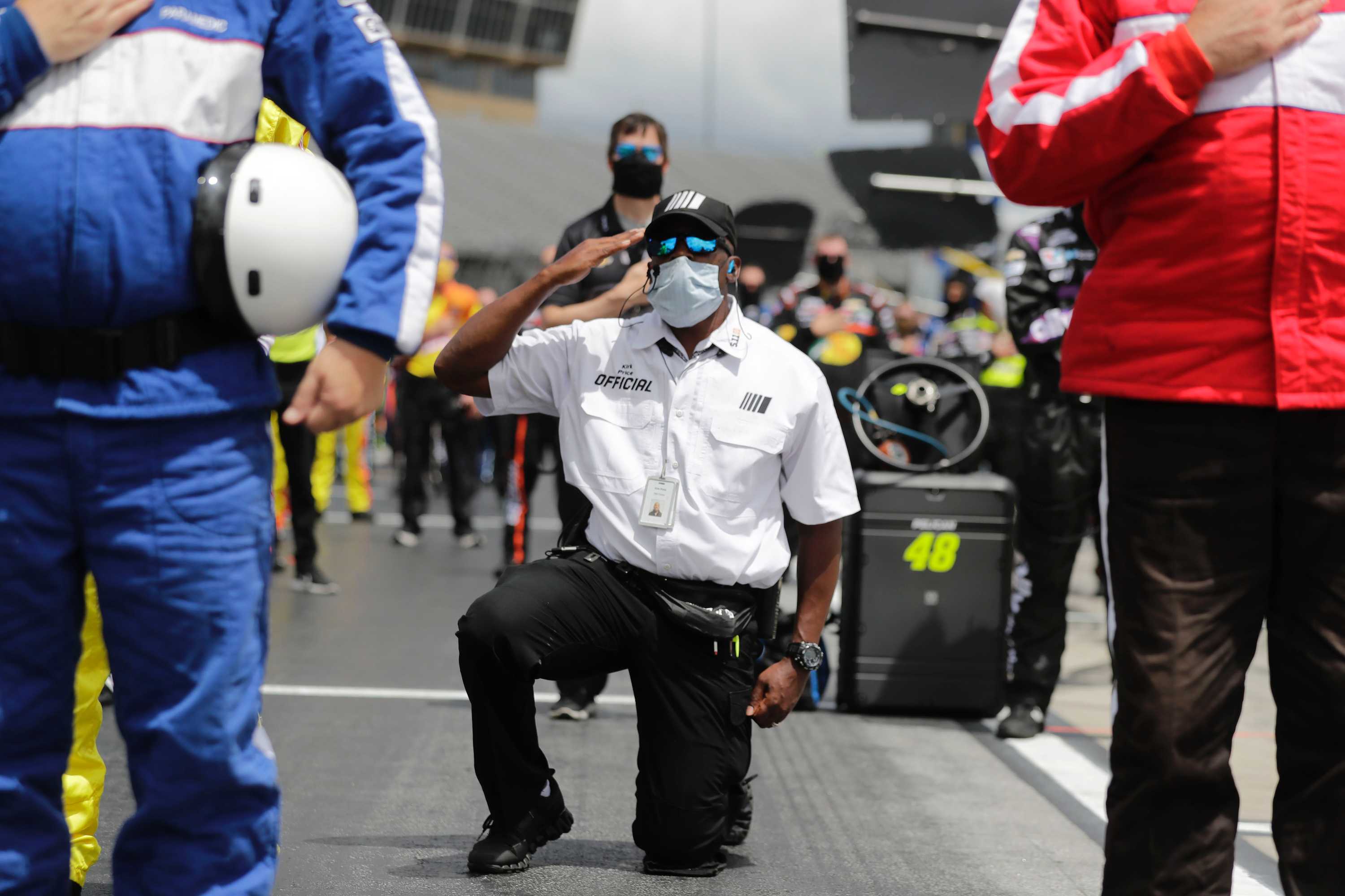 An official kneels while saluting the flag (not visible) during the national anthem at a NASCAR race.