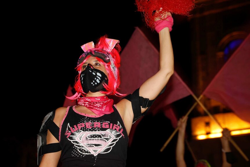 A woman wearing a black mask and pink headgear and a pink bandana around her neck holder her arm in the air