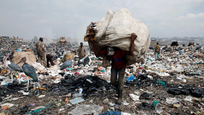 A scaveger carries a large sack over the bag of his head through a dumping site.
