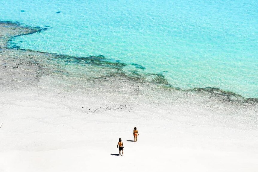 A drone photo of two people on a white beach with bright blue ocean.