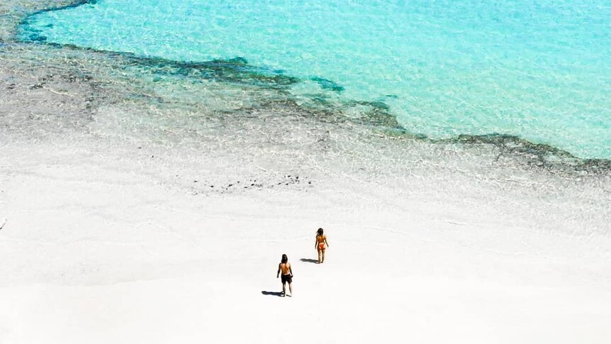 A drone photo of two people on a white beach with bright blue ocean.