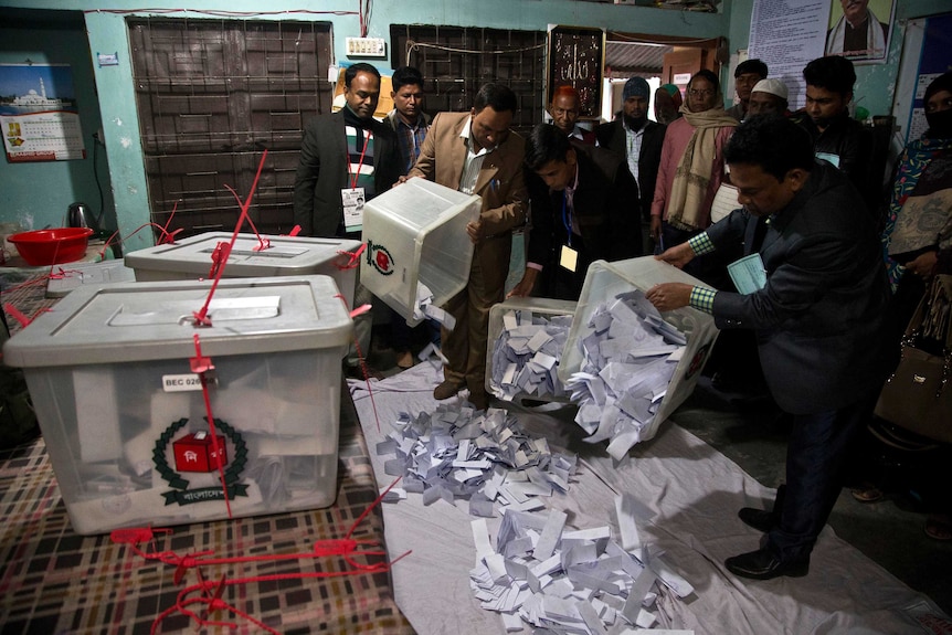 Officials wearing lanyards empty plastic tubs full of ballot papers onto the ground inside a packed room.