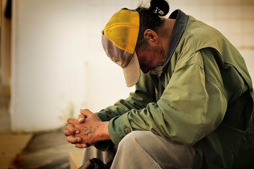 A tatooed homeless man with his head down. He's wearing a green long-sleeved shirt and trousers as well as a yellow cap.