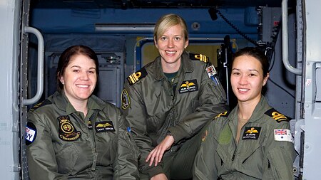Four women in flying suits stand in front of a helicopter.