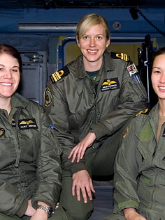 Four women in flying suits stand in front of a helicopter.